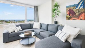 A modern living room with a grey sectional sofa, a small round coffee table, and a view of the ocean through large windows. - Excelsior I Casa LEX Ocean&Sunrise Views Steps to Beach&Dunes Terrace Pool (Playa del Ingles)