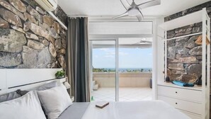 A bedroom with a stone wall, a bed with grey bedding, a ceiling fan, and a view of the sea through a sliding door. - Excelsior I Casa LEX Ocean&Sunrise Views Steps to Beach&Dunes Terrace Pool (Playa del Ingles)