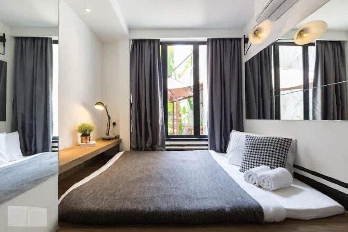 A modern hotel room with a large bed, a bedside table with a lamp, and a view of the outdoors through a sliding door.