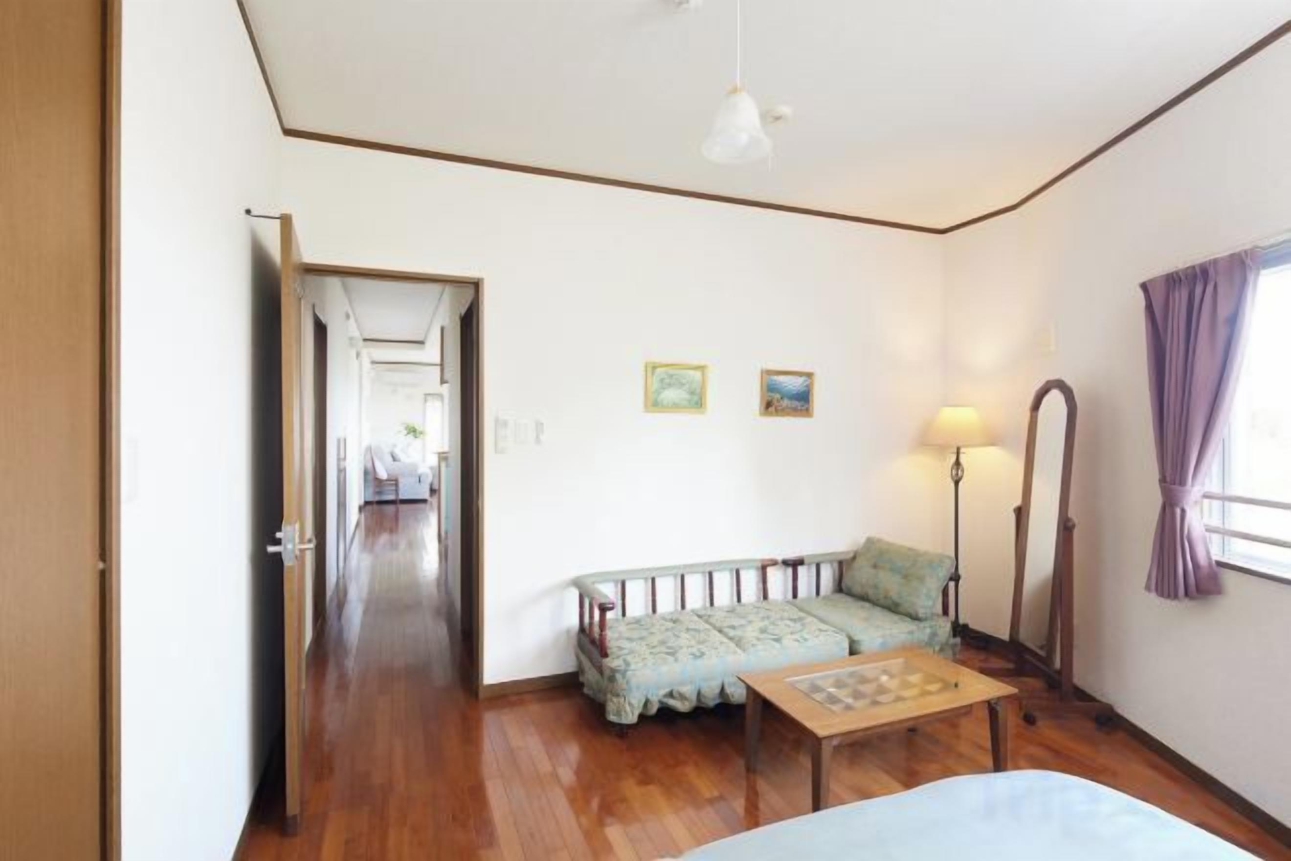 A room with a wooden floor, a sofa, a small table, a lamp, and a bed. There is a door leading to another room.