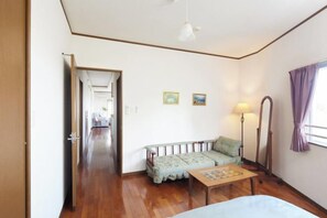 A room with a wooden floor, a sofa, a small table, a lamp, and a bed. There is a door leading to another room. - Ocean Therapy House (Onna)