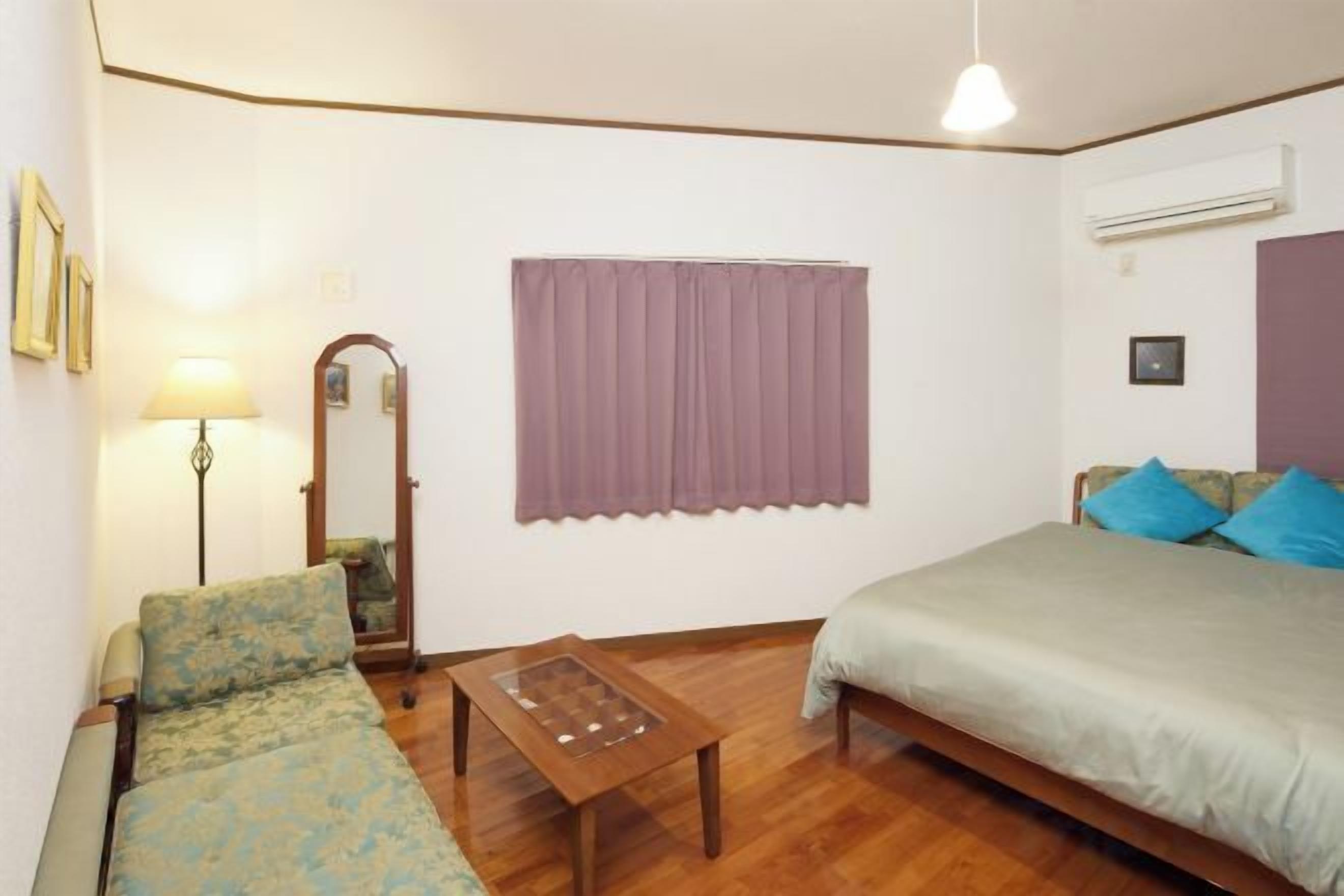 A room with a bed, a sofa, a table, a lamp, a mirror, and a window with curtains.