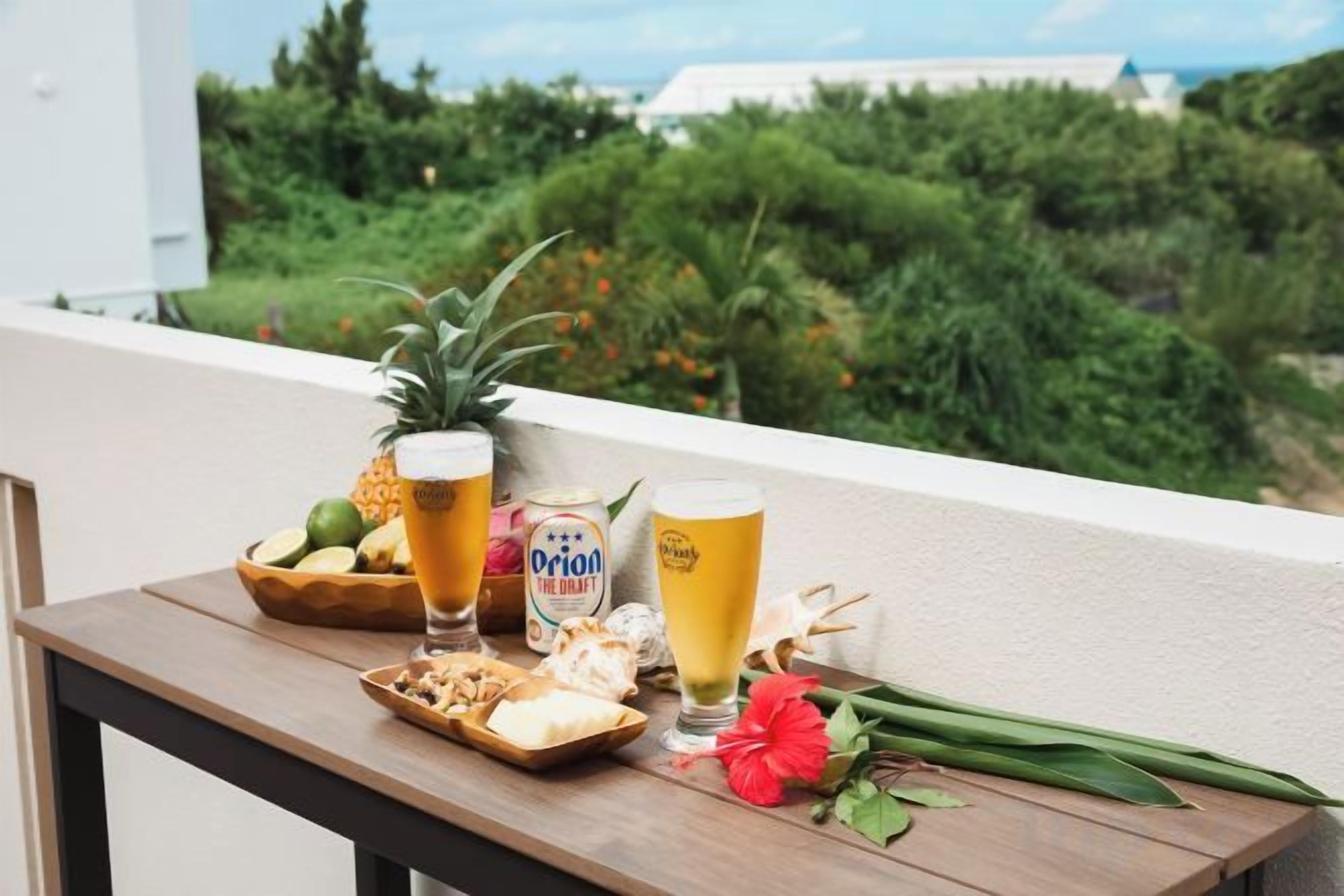 A table with drinks, a pineapple, a bowl of snacks, and a flower on a balcony with a view of greenery and a building.