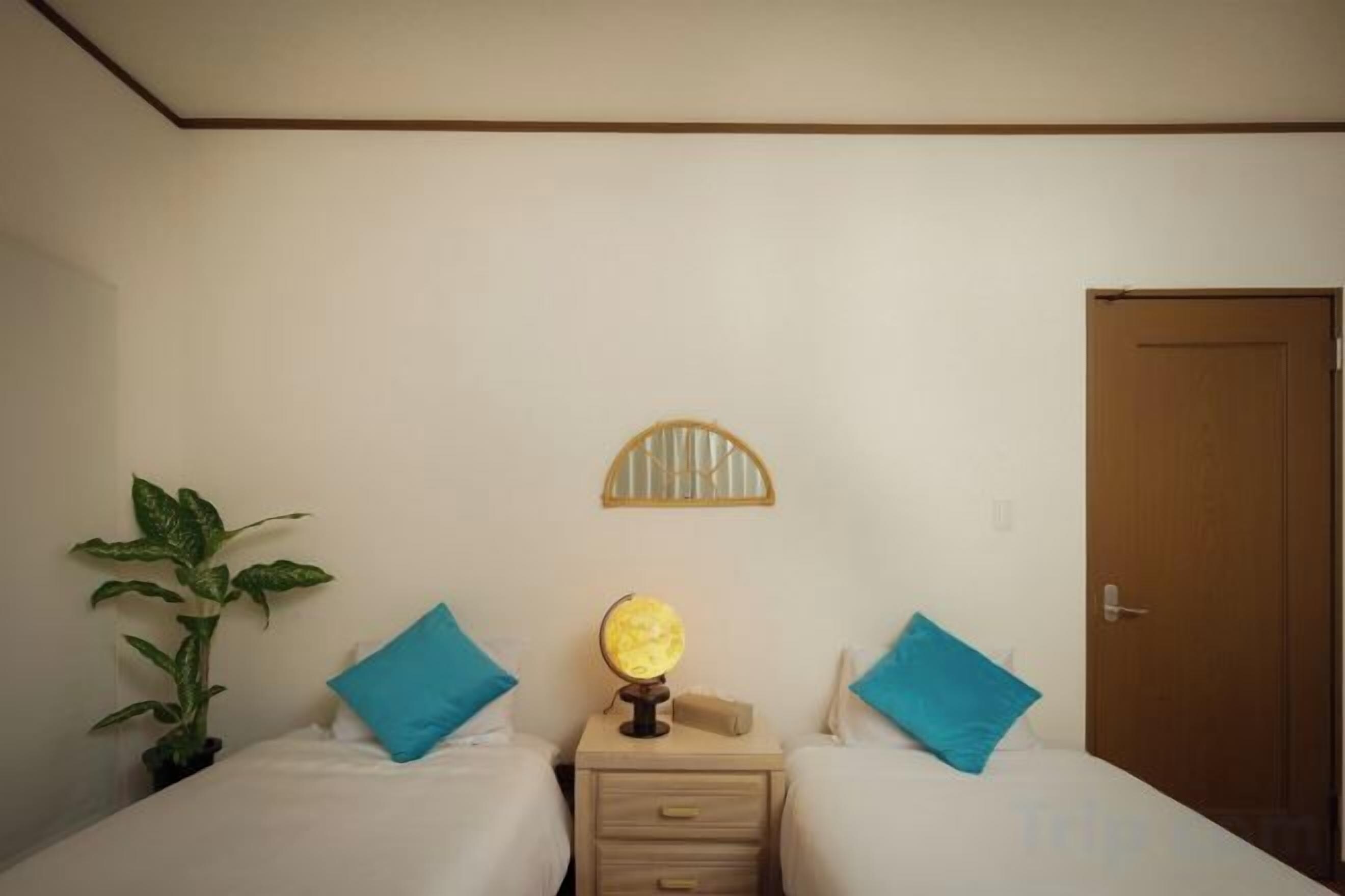 Een kamer met twee bedden, een nachtkastje met een lamp, een plant in pot en een deur.