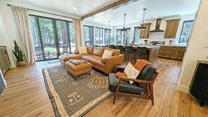 A spacious living room with a large sofa, a coffee table, and a rug. There is a kitchen area with bar stools and a dining table. - Pine Mountain 6 - BRAND NEW HOME! 4 Bd, 4 Bath, SHARC, Hot Tub + More! (Sunriver)
