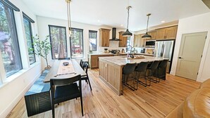 A modern kitchen with large windows, wooden cabinets, and a central island with bar stools. - Pine Mountain 6 - BRAND NEW HOME! 4 Bd, 4 Bath, SHARC, Hot Tub + More! (Sunriver)