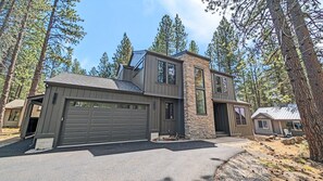 Une maison moderne à deux étages avec un garage, entourée de grands pins.