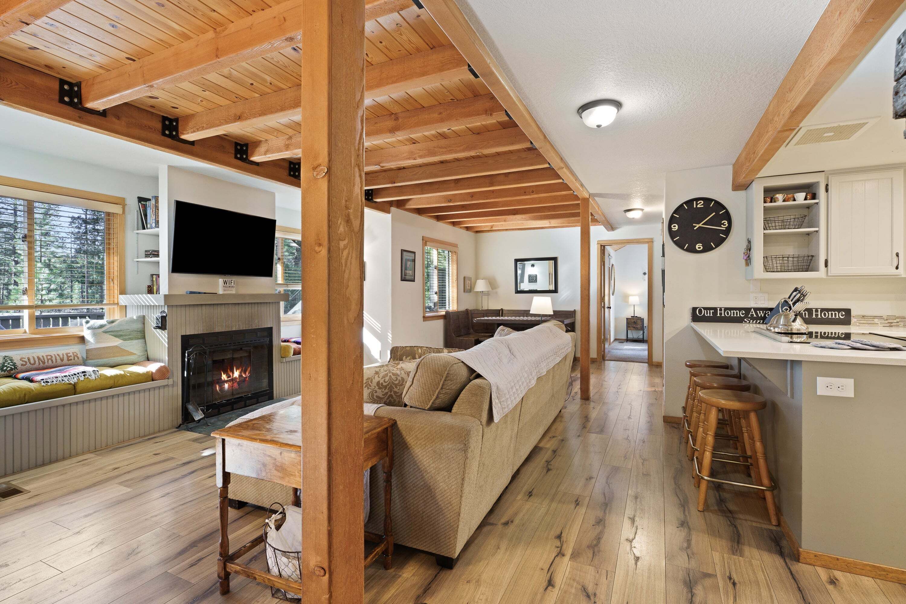 A cozy living area with a fireplace, wooden beams, a sofa, and a kitchen area with a clock and bar stools.