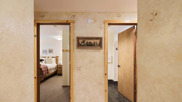 A hallway with wooden doors leading to a bedroom with a bed, a nightstand, and a wall-mounted picture.