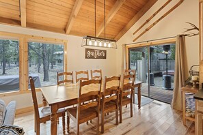 A dining area with a wooden table and chairs, a sign reading 'gaTher,' and a view of the outdoors through large windows. - Pine Needle 2 - A/C, Hot Tub, SHARC (Sunriver)