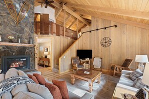 A spacious living room with a stone fireplace, wooden ceiling beams, a staircase, and a large wall clock. - Pine Needle 2 - A/C, Hot Tub, SHARC (Sunriver)