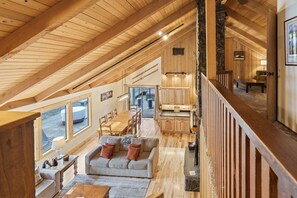 A spacious loft with a high ceiling, wooden beams, a dining area with a long table and chairs, a kitchen with wooden cabinets, and large windows offering views of the outdoors. - Pine Needle 2 - A/C, Hot Tub, SHARC (Sunriver)