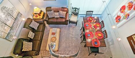 Un coin repas avec une longue table dressée pour six personnes, entourée de chaises. On y trouve également un canapé avec des coussins à motifs et une table basse agrémentée d’objets décoratifs.