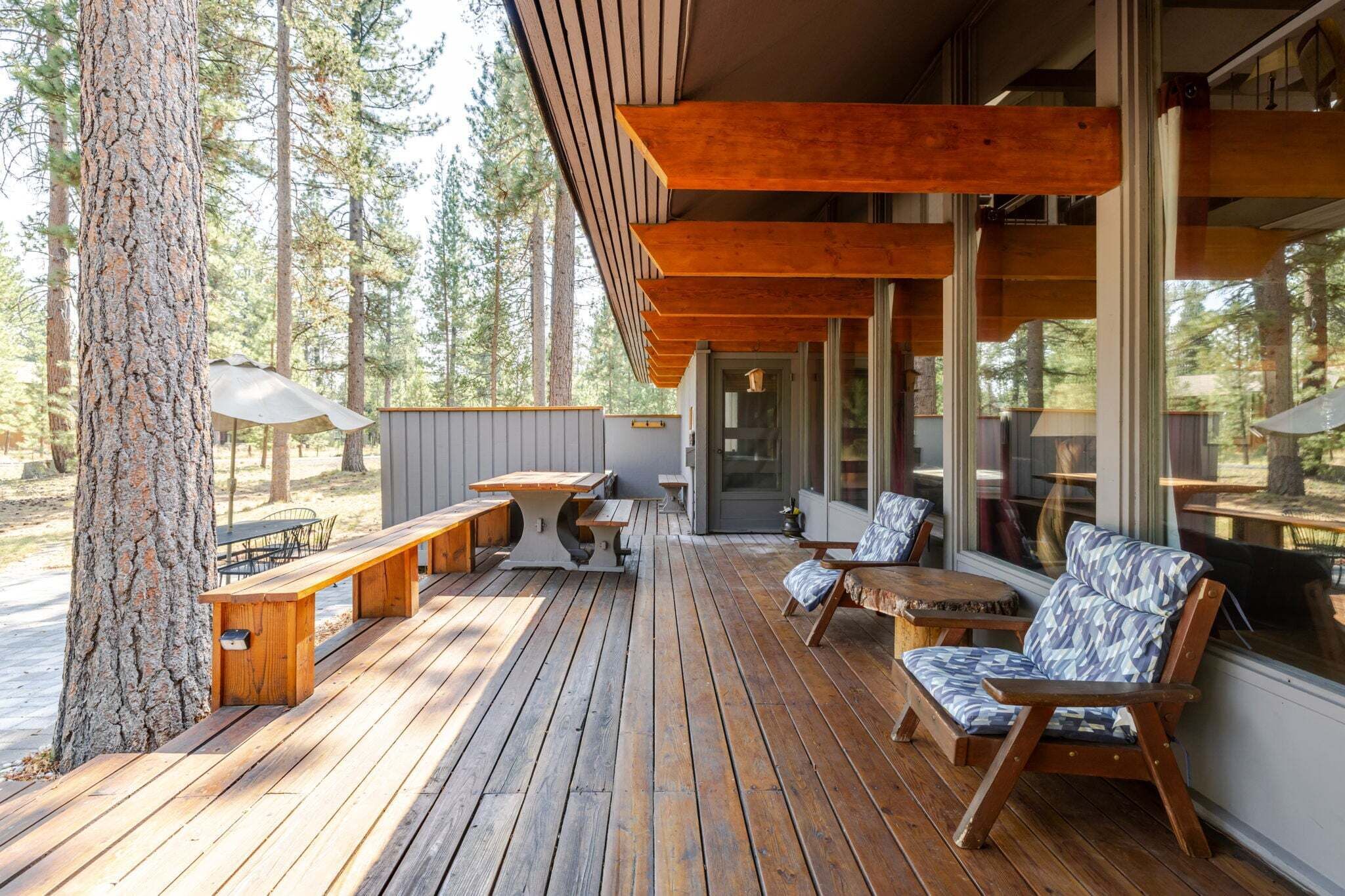 Une terrasse en bois avec des sièges d’extérieur, de grandes fenêtres et une vue sur les arbres.