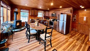 A cabin interior with a wooden floor, a round dining table, chairs, a kitchen area with a stove, microwave, and refrigerator, and a large window. - The Perfect Adirondack Getaway Waterfront Cabin (Northville)