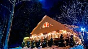 A snow-covered house with a lit balcony and snow-covered trees. - The Perfect Adirondack Getaway Waterfront Cabin (Northville)