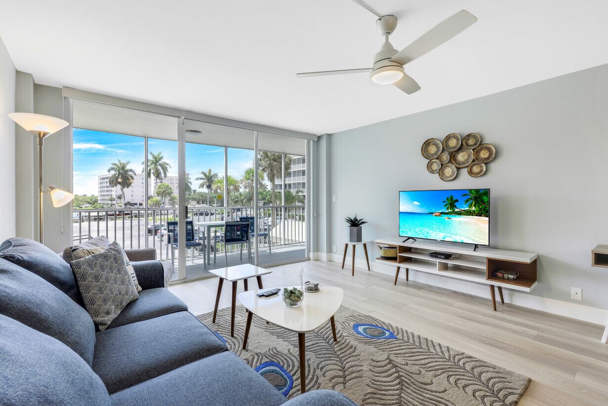 A modern living room with a large TV, grey sofa, and a balcony with a view of palm trees and buildings.