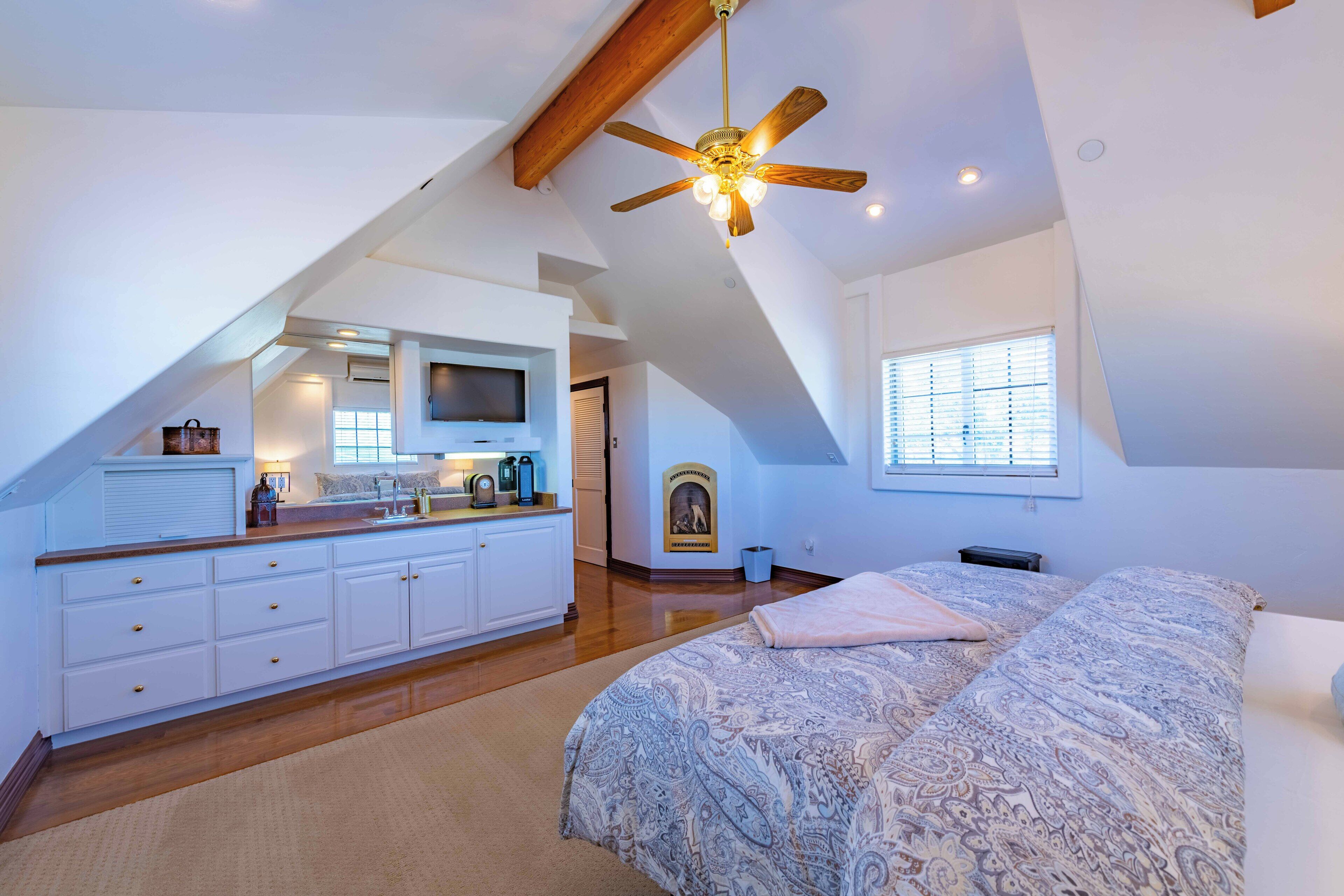 Un dormitorio con una cama grande, un ventilador de techo, televisión y una zona de cocina con armarios y ventana.