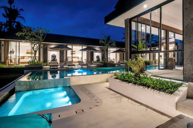 A modern outdoor pool area with lounge chairs and umbrellas, surrounded by a well-maintained garden and a contemporary building with large windows.