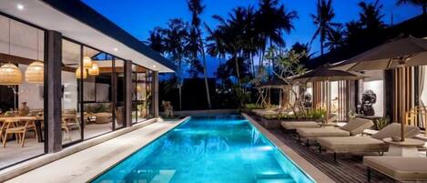 A swimming pool with lounge chairs, a glass-enclosed room, and palm trees in the background.