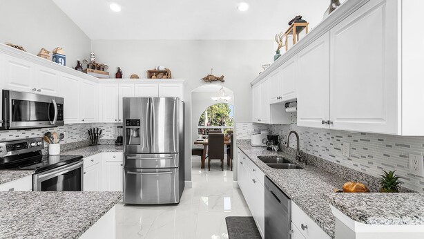 A modern kitchen with white cabinetry, stainless steel appliances, granite countertops, and a view to the dining area. - Dive into Endless Summer Bliss - Forever Summer - Roelens (Cape Coral)