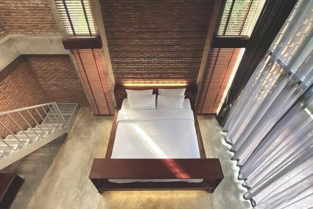 A high-ceilinged room with a bed centered, a staircase to the left, and large windows on the right.