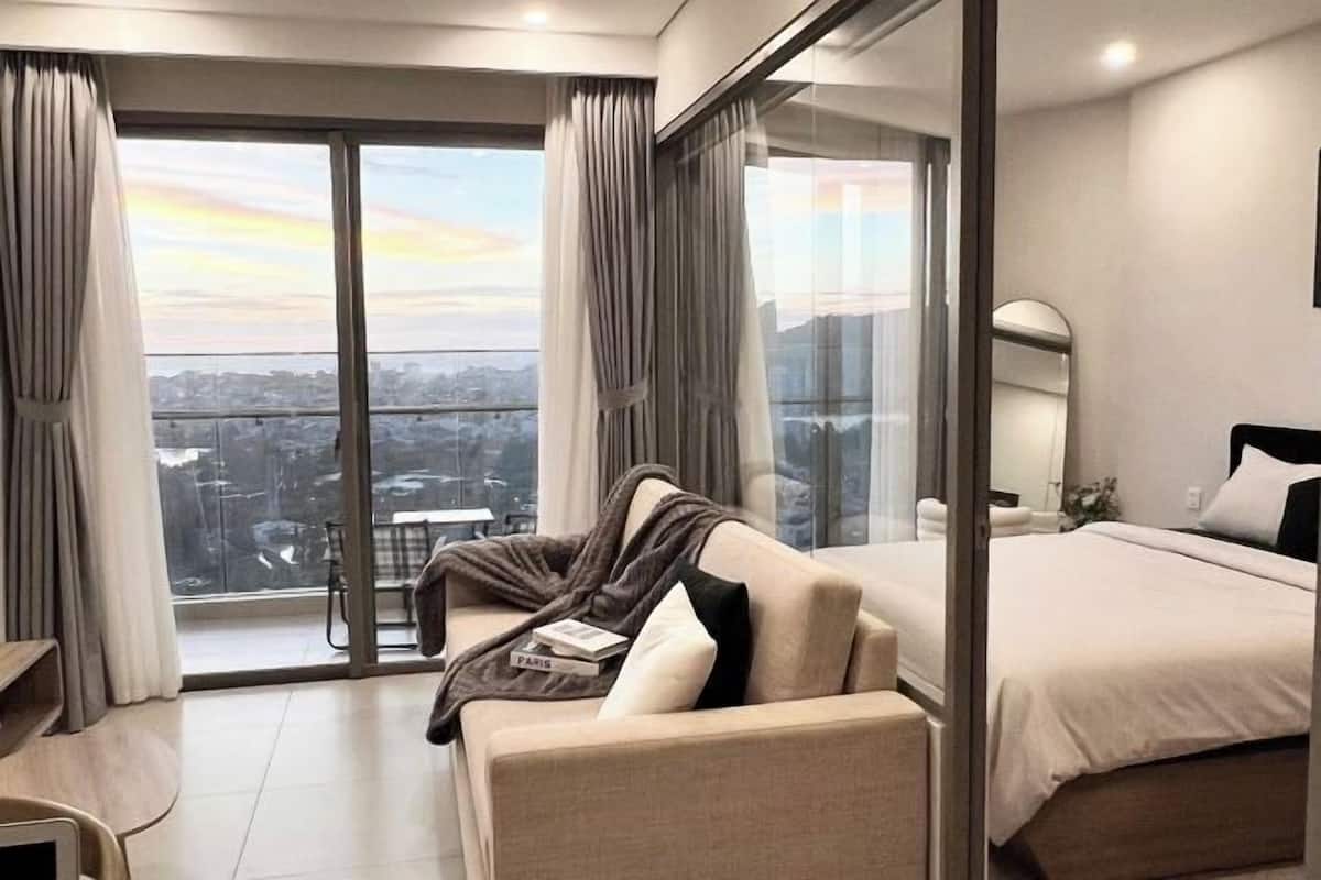 Quarto de hotel moderno com uma cama grande, um sofá, porta de vidro para uma varanda e vista para a cidade.