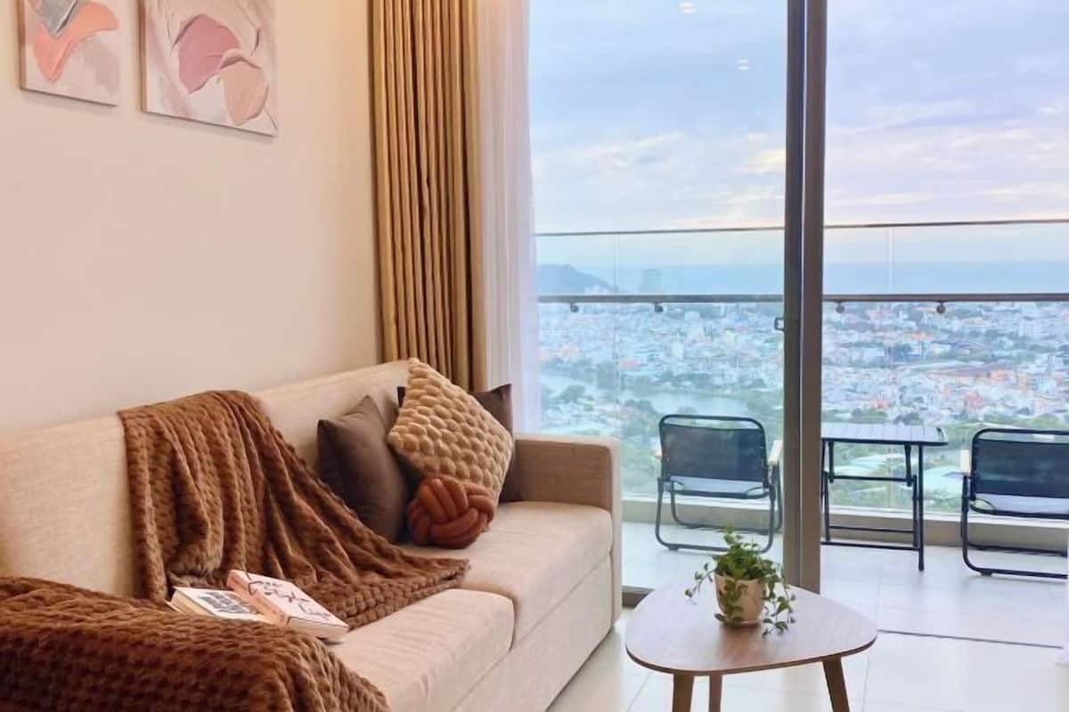 Sala de estar moderna com sofá, mesa de centro e vista para o horizonte urbano através de grandes janelas.