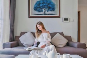A person sitting on a sofa with a book, a coffee table with a teapot and cups, and a painting of a tree on the wall. - Landmark Cityview Apartment (Ho Chi Minh City)
