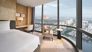 A hotel room with a large bed, a desk, a chair, and a view of the city through large windows.