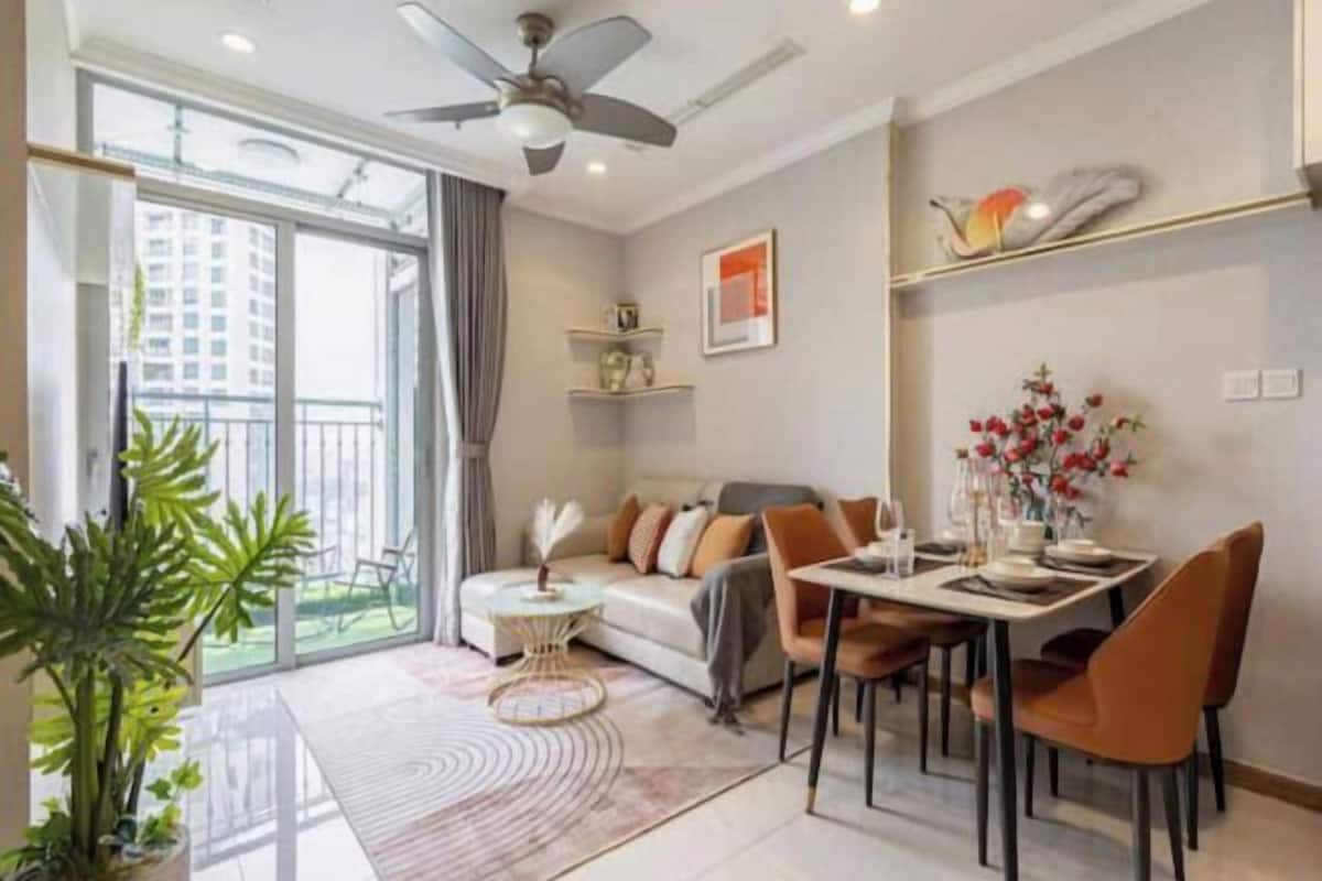 A modern living room with a sofa, dining table, chairs, a ceiling fan, and a large window with a view of buildings.