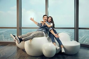 A couple sitting on a large, white, bean-shaped chair in a room with large windows offering a city view. - Landmark Cityview Apartment (Ho Chi Minh City)