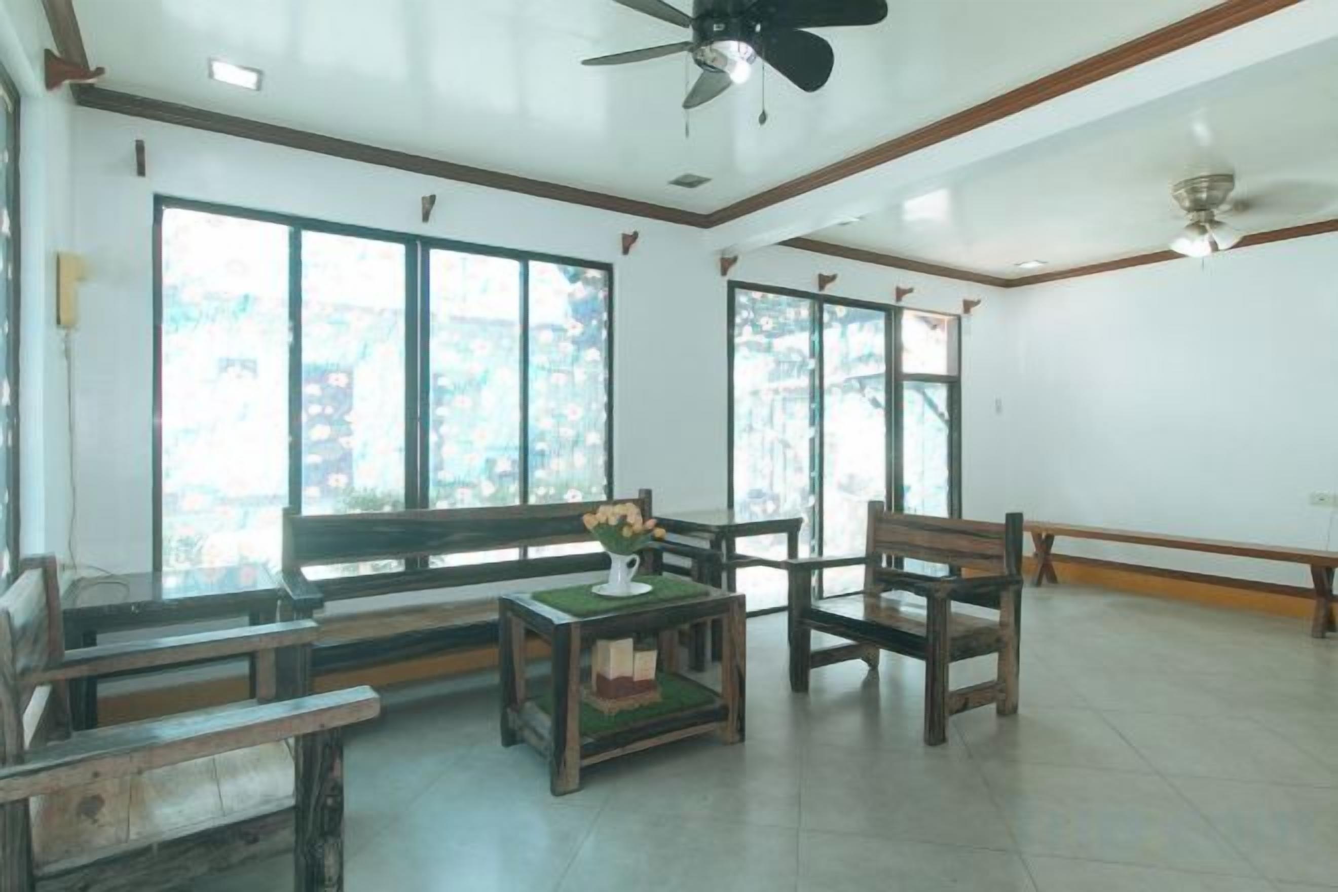 A spacious room with a high ceiling, large windows, and a ceiling fan. There are wooden benches and a small table with a vase of flowers.