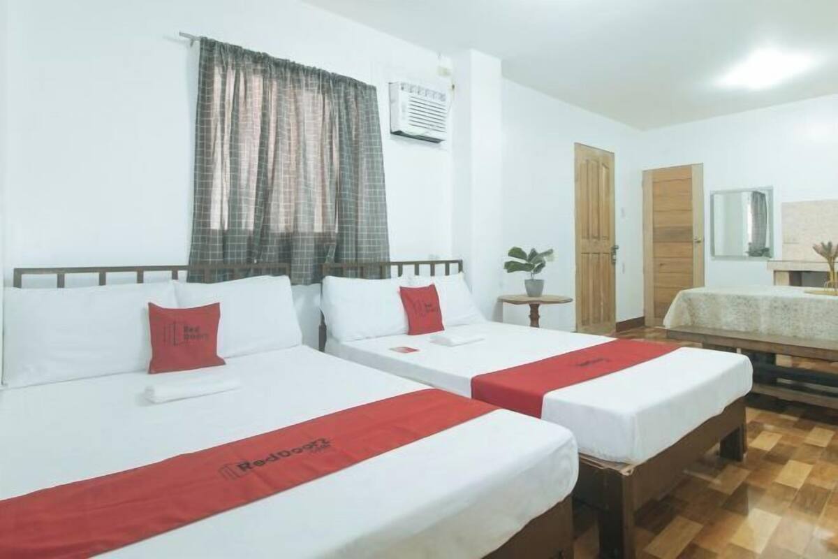 A hotel room with two beds, a wooden bench, a small table, a mirror, and a window with curtains.