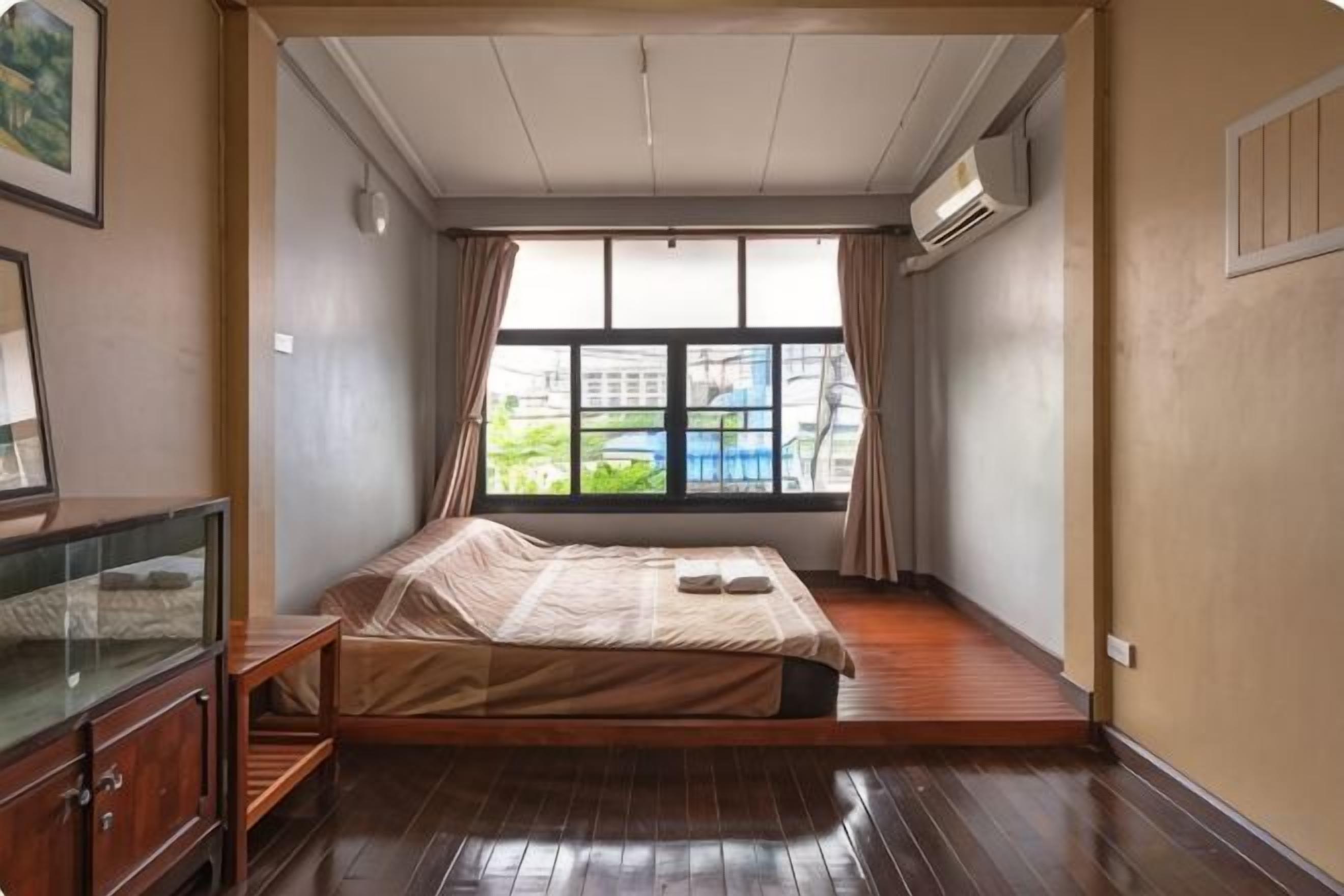 A room with a bed, a wooden floor, a window with curtains, and a view of buildings outside.