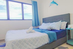 Una cama con colcha azul y blanca, una mesita de noche con lámpara, y una ventana con cortinas.