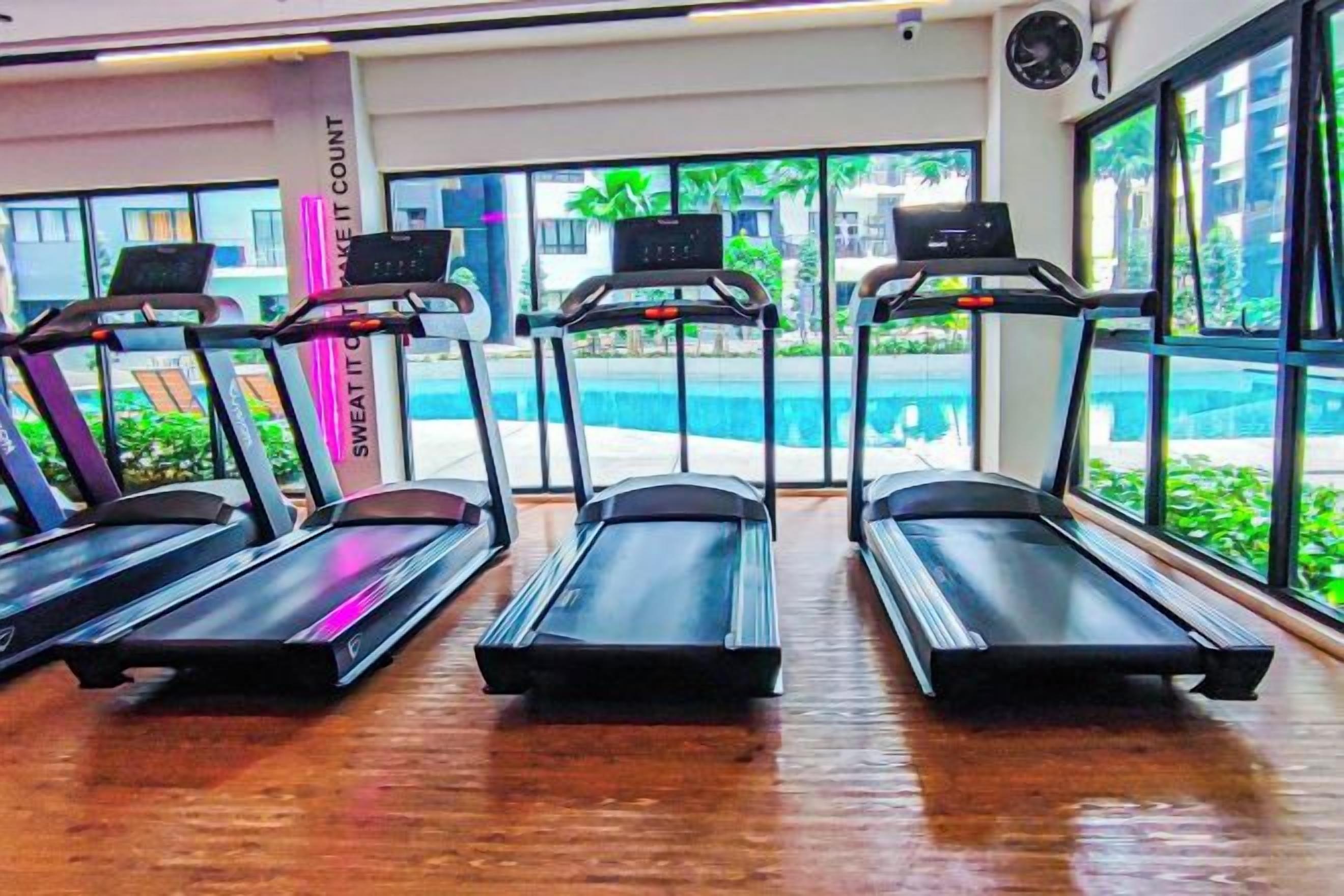 A gym with treadmills aligned in a row, large windows, and a view of a pool outside.