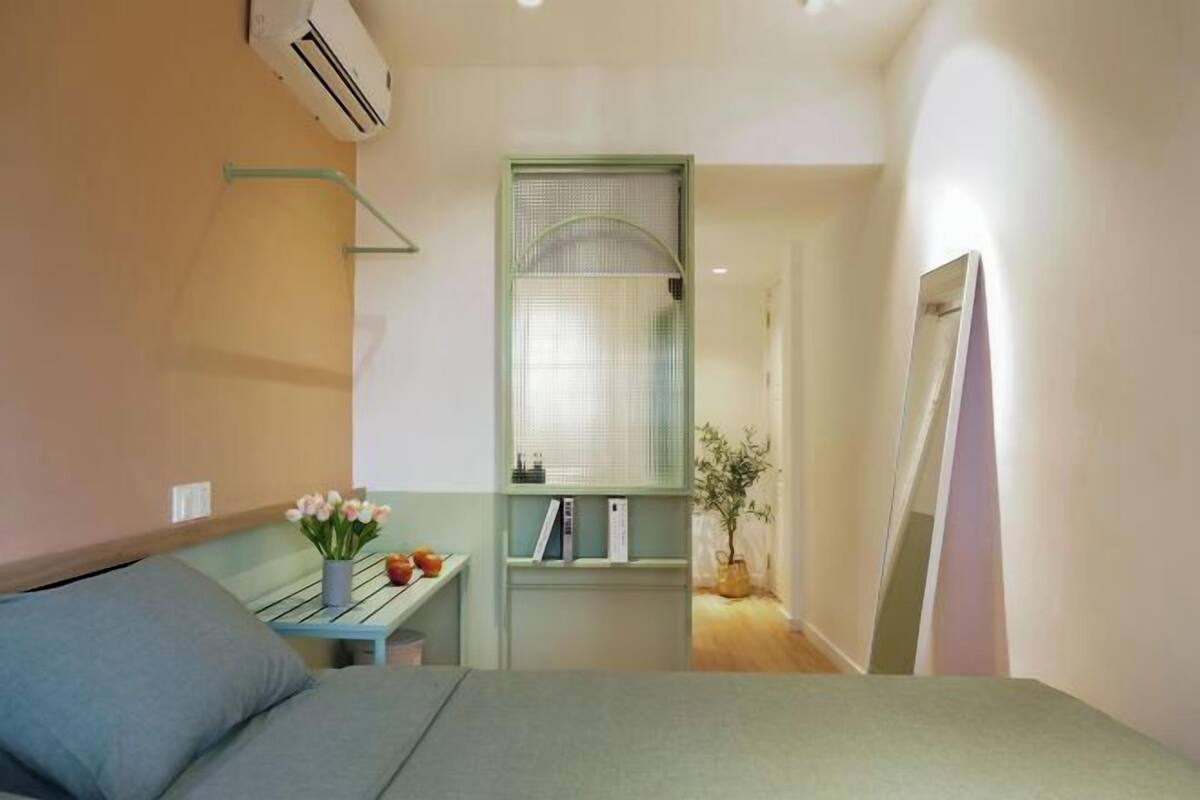 Une chambre à coucher avec un lit, une petite table sur laquelle est posé un vase de fleurs, une fenêtre avec des stores et un miroir au mur.