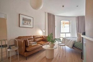 A modern hotel room with a leather sofa, a small round table with a plant, a bed with a green bedspread, and a framed picture on the wall.