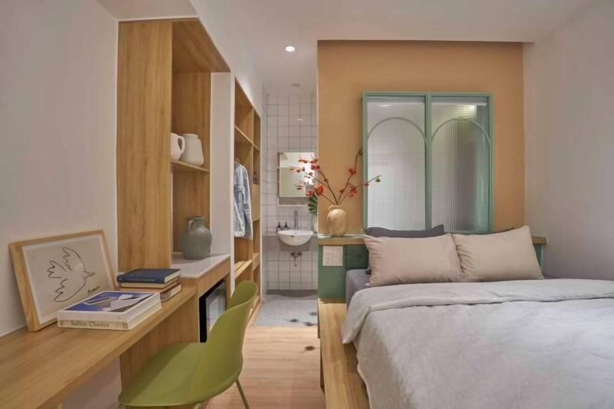 Une chambre à coucher comprenant un lit, un bureau avec une chaise, une bibliothèque, un miroir et une salle de bain avec un lavabo et des toilettes.