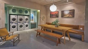 A laundry room with multiple washing machines, a wooden bench, a table, and two chairs. - Express by M Village Pham Ngu Lao, a brand of Modern Village Lifestyle (Ho Chi Minh City)
