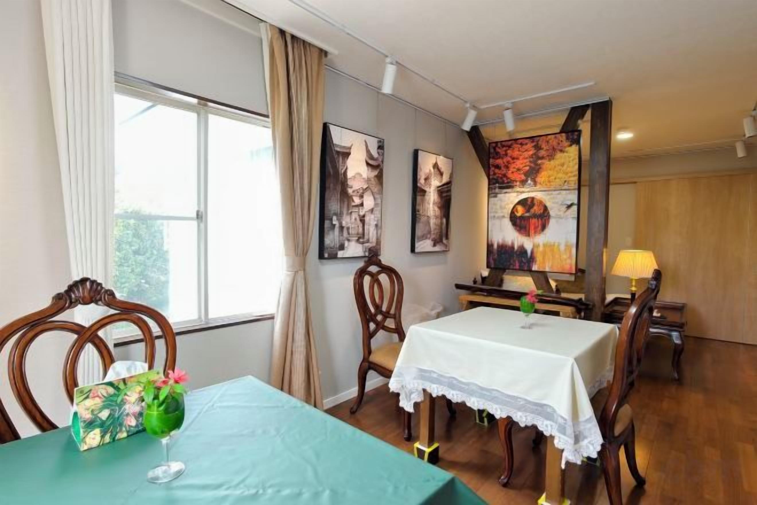 Un coin repas avec une table dressée pour deux personnes, des chaises en bois, un vase avec des fleurs et une fenêtre avec des rideaux.