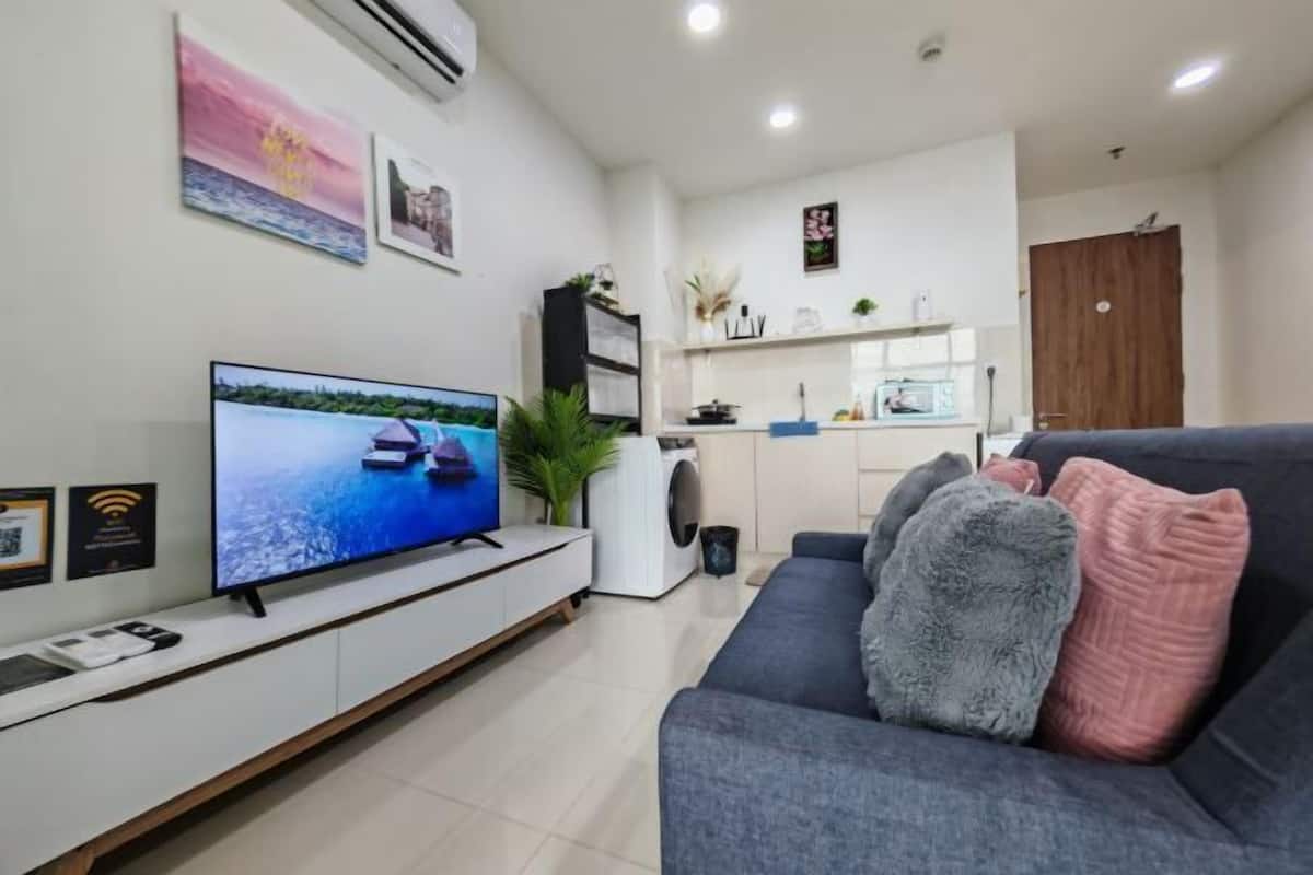 A modern living room with a flat-screen TV, a grey sofa, and a kitchen area in the background.