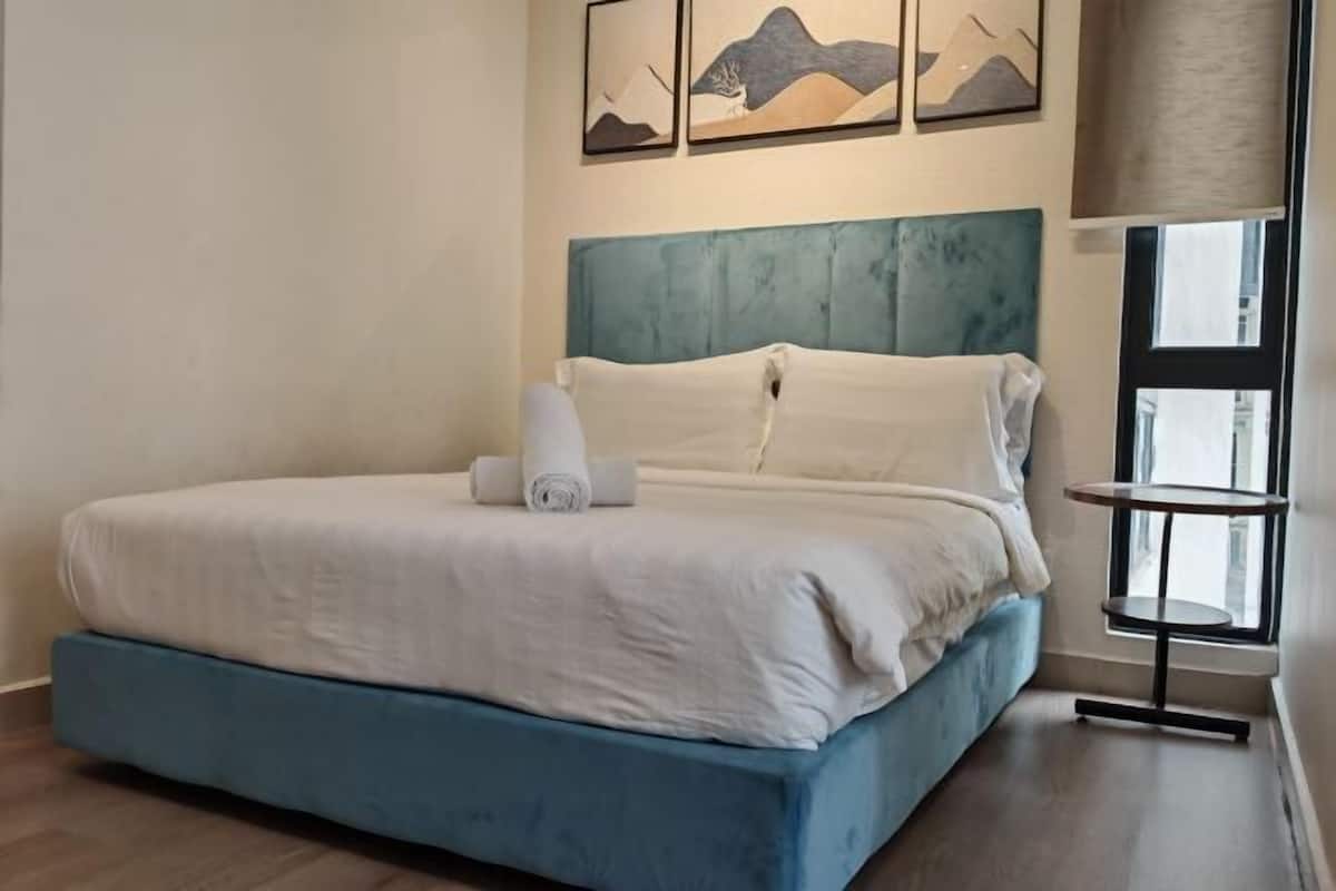 A neatly made bed with a blue headboard and white bedding, a small bedside table, and artwork on the wall.