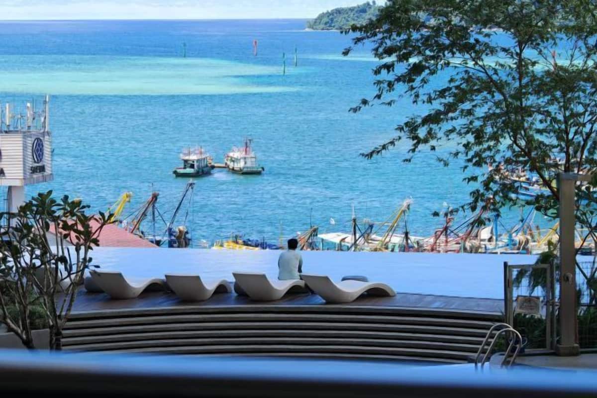 A pool area with a view of the sea and boats, a lifeguard tower, and a person sitting on a lounge chair.