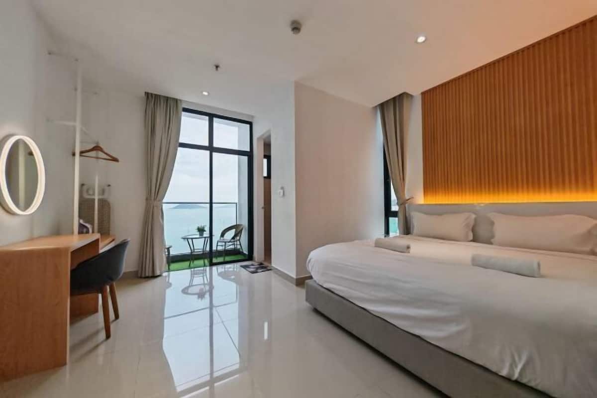 A modern hotel room with a large bed, a desk, a chair, a mirror, and a view of the sea through a large window.