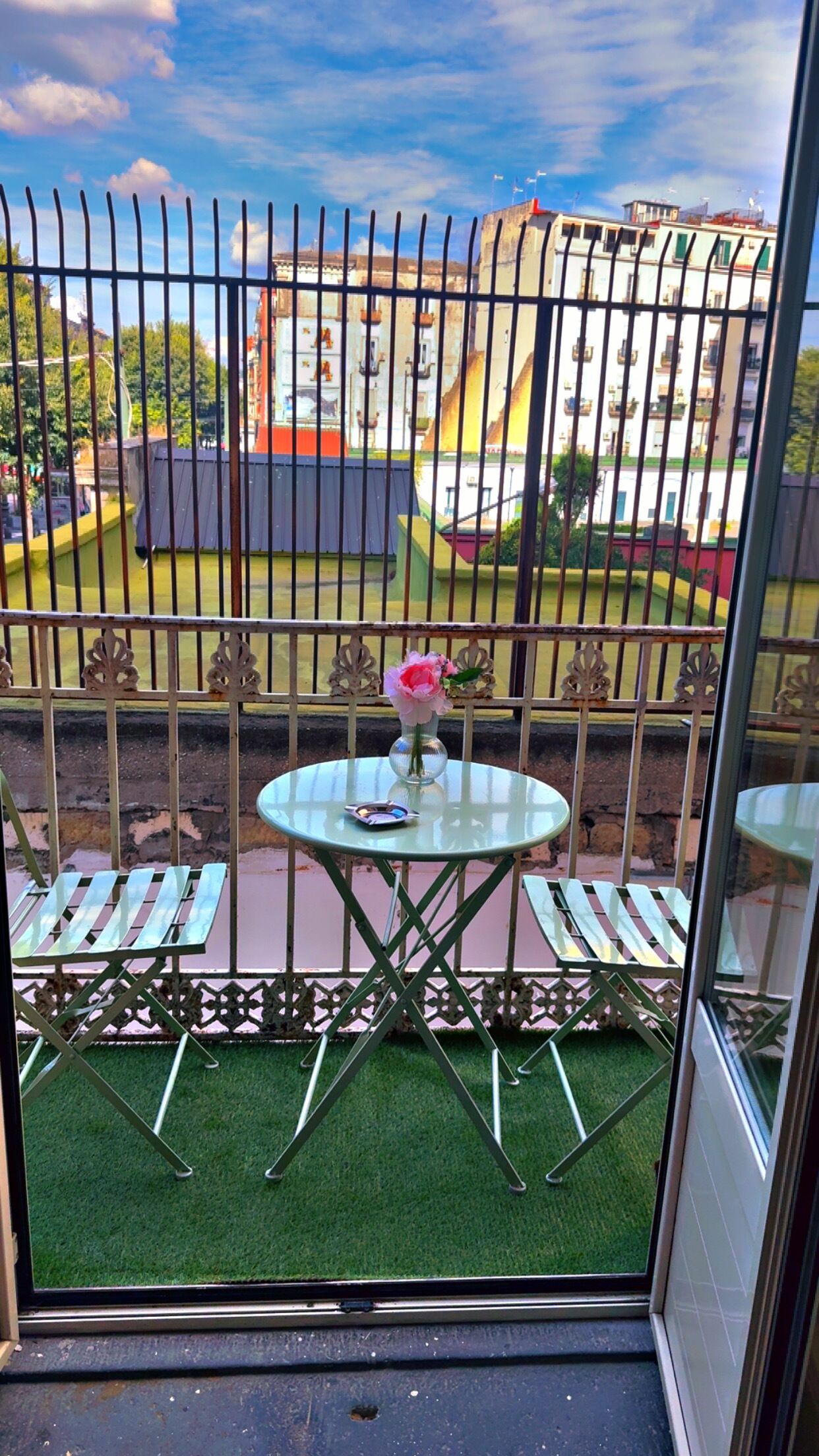 Ein Balkon mit kleinem Tisch und Stühlen, einer Blumenvase und Blick auf Gebäude und Grünflächen.