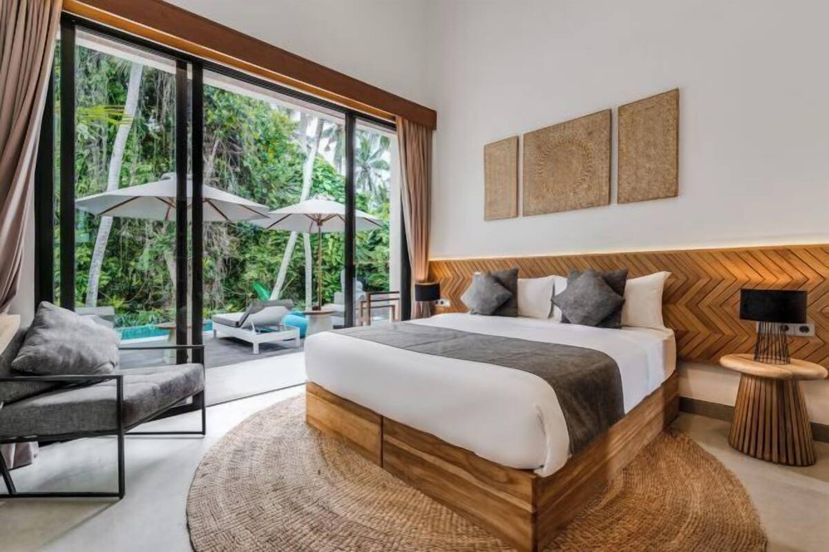 A modern bedroom with a large bed, a chair, a nightstand, and a view of a pool and greenery.