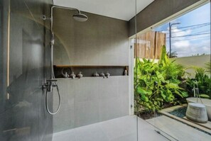 A modern shower area with a glass door, a built-in shelf with decorative items, and a view of a lush garden outside. - Villa Moana by BaliSuperHost (Bali)