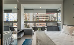 A hotel room with a large bed, a desk, two chairs, and a view of the cityscape through large windows. - Studio Premium Oscar Freire (São Paulo)